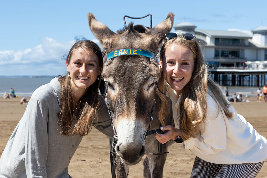 Two ladies standing close either side of a donkey they are cuddling